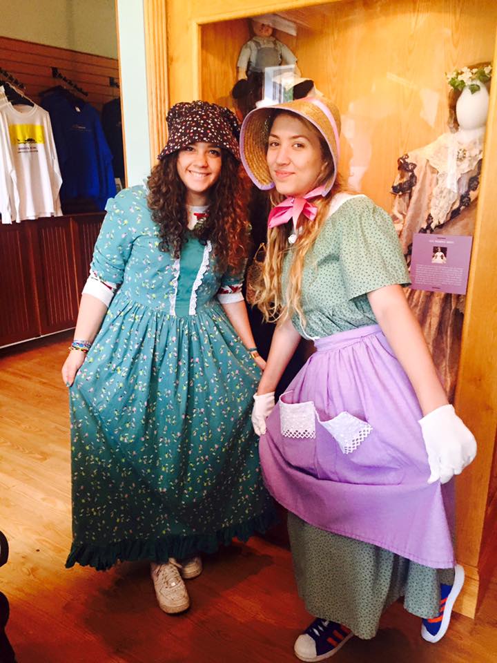 Two girls posing wearing pioneer outfits. They are standing in front of a display cabinet featuring a mannequin wearing another outfit.