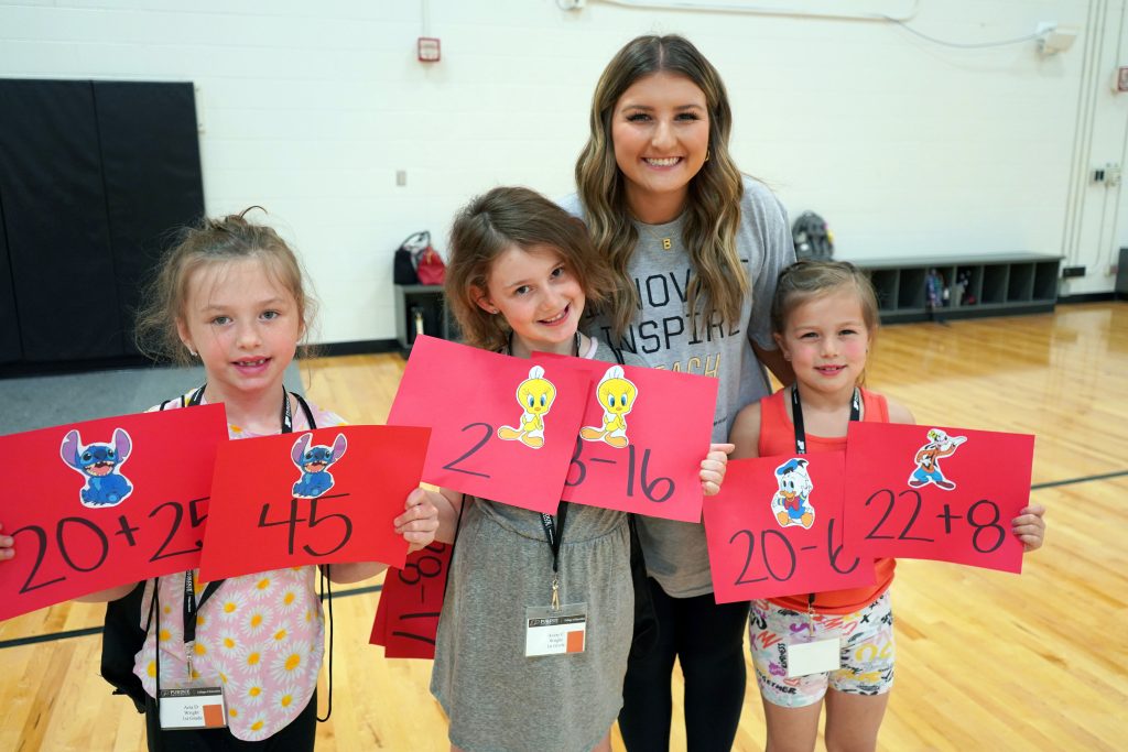 A Purdue College of Education student smiling for the camera with three schoolchildren. The three girls are holding up pieces of construction paper with math problems on them.