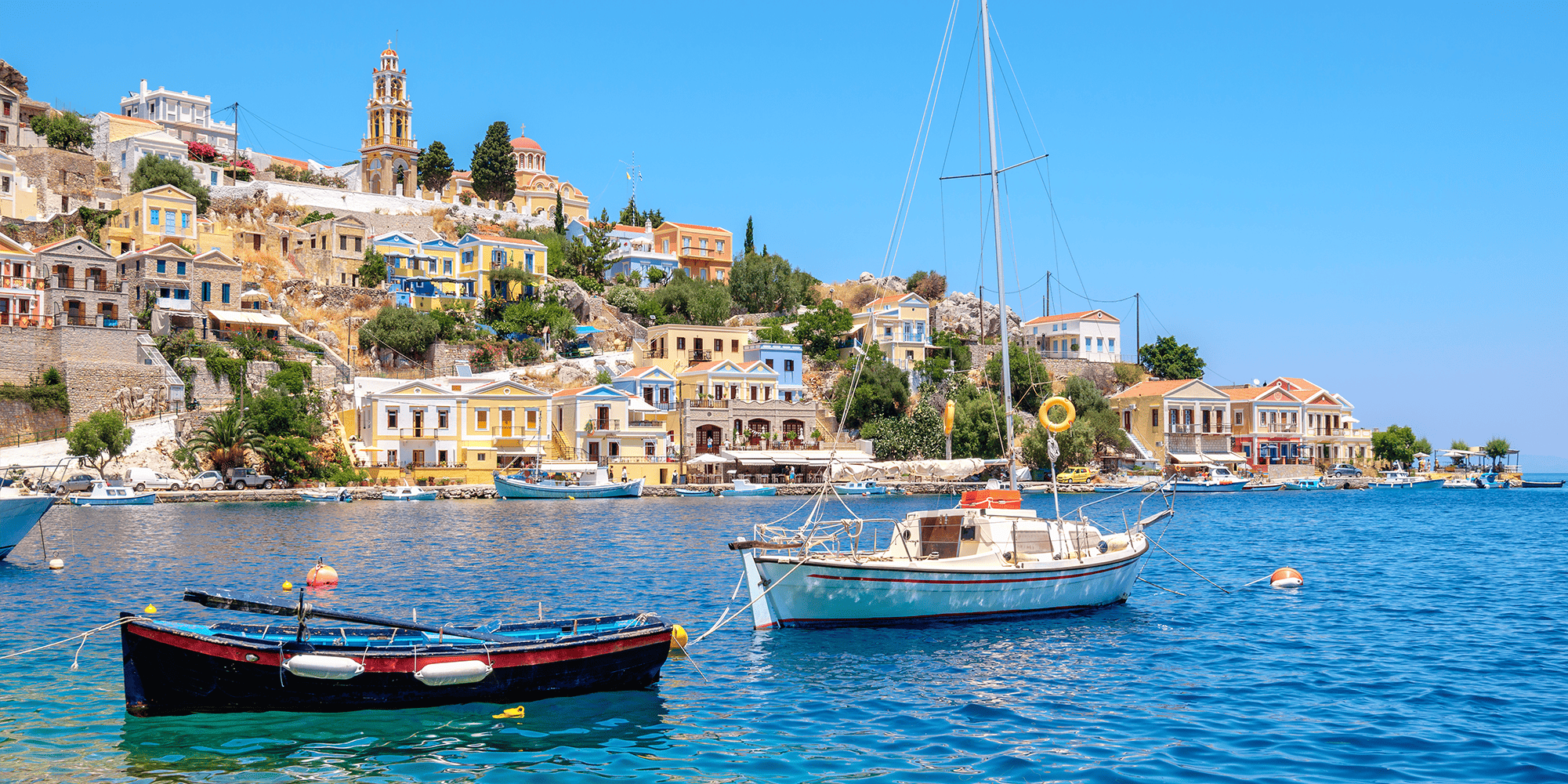 A view of Greece's coastline. In the foreground are two boats in an ocean. In the background is colorful Greek architecture. 