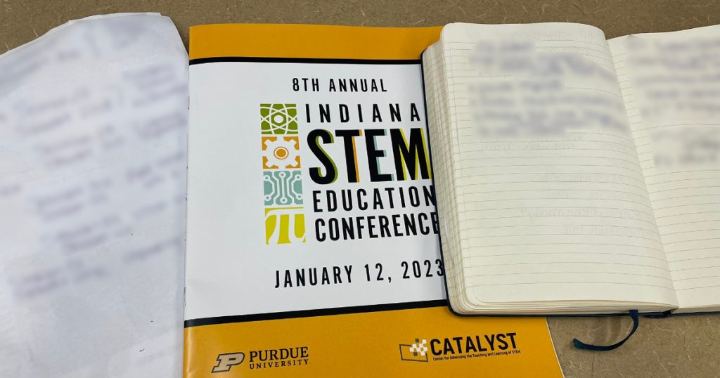 Two notebooks with a white and gold folder between them with the following text: 8th Annual  Indiana STEM Education Conference January 12, 2023