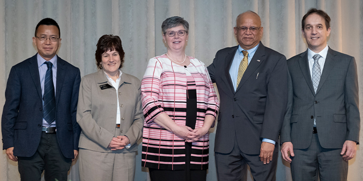 Kristen Seward, Nielsen Pereira, and three other faculty standing at a podium in front of a grey backdrop.