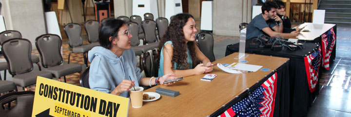 Two students sitting at a table smiling at someone off screen. On the table is a sign with "Constitution Day" on it and a banner with an American flag pattern.
