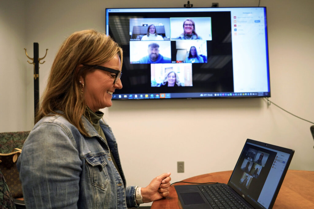 Side view of Alice Johnson smiling at a computer in a virtual meeting with graduate students. Behind her is a large screen mirroring the meeting.