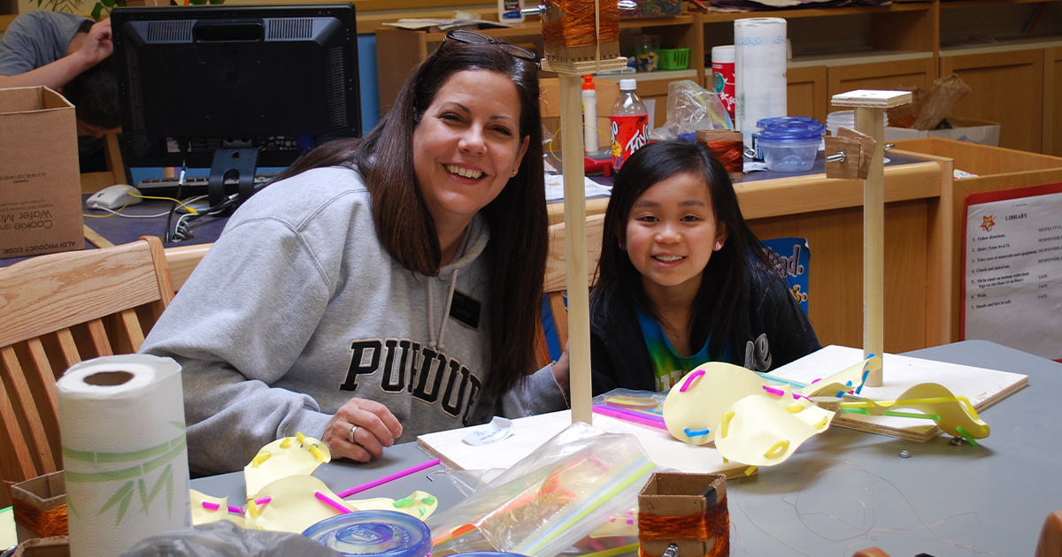 Lynn Bryan, executive director of CATALYST, and a student seated at a table and smiling for a photo. On the table in front of them is a wooden wind turbine model.