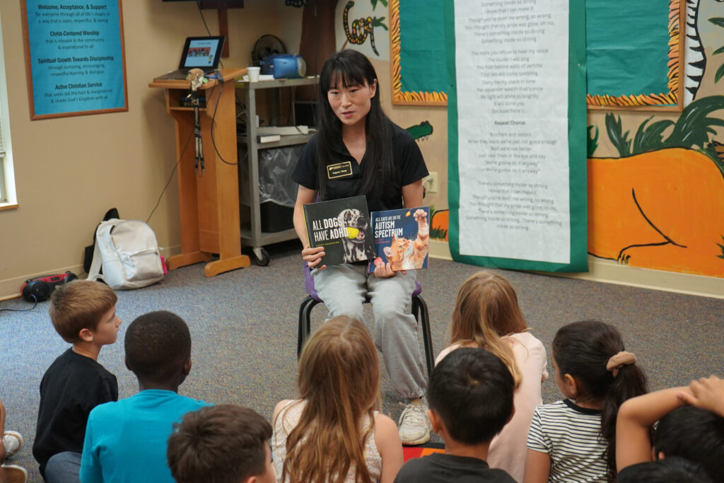 Jingyuan Zhang teaching about disabilities at the Purdue CDF Freedom School® Program. She is seated in a chair in front of a group of children who are seated on the ground. In her hands are two books titled "All Dogs have ADHD" and "All Cats are on the Autism Spectrum".