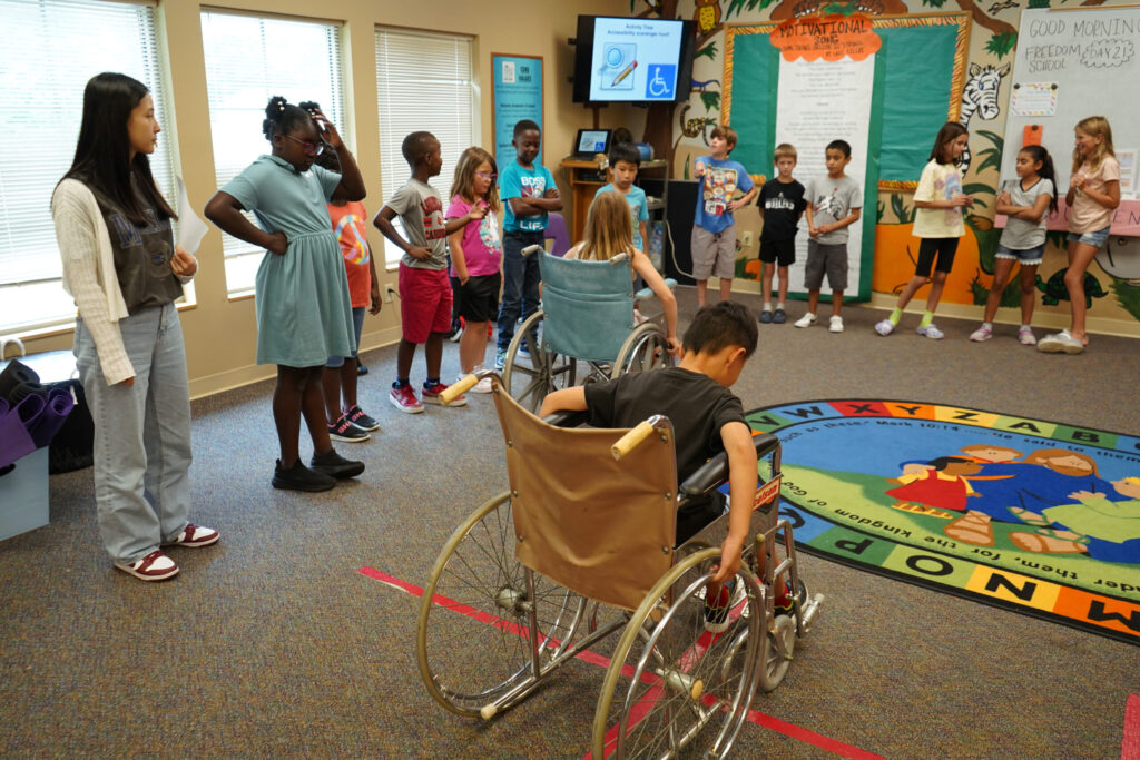 Participants at the Purdue CDF Freedom School® Program explore what it might be like 
to use a wheelchair. Two students are seated in blue and yellow wheelchairs. A crowd of students are standing around them.