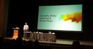Presenter Phil Cook standing on an auditorium stage. Behind him is a presentation titled "Curiosity, Sticky Learning & Social Media"