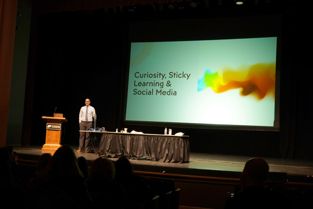 Presenter Phil Cook standing on an auditorium stage. Behind him is a presentation titled "Curiosity, Sticky Learning & Social Media"