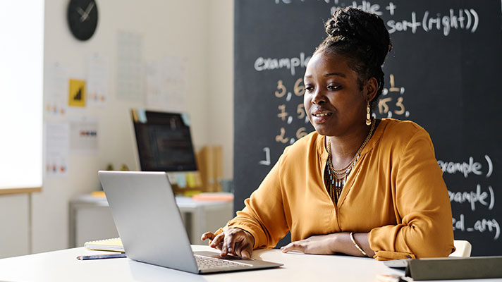 A black female works on a laptop in front of a blackboard in a classroom