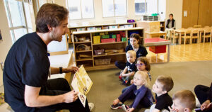 A graduate student reading a book to a small group of children, who are seated on the floor.