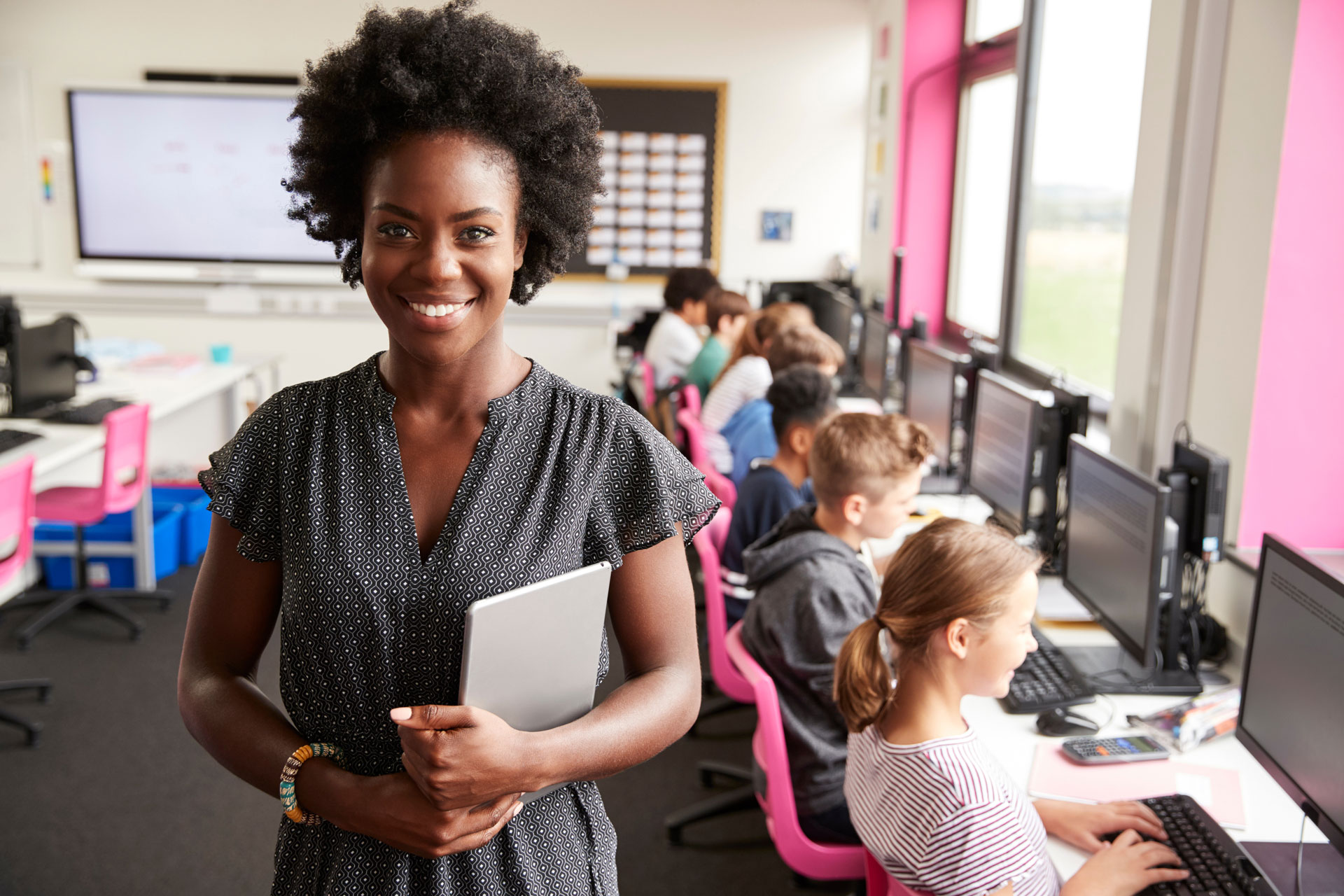 A black female teacher stands in front of a classroom. Students are on computers in the background. 