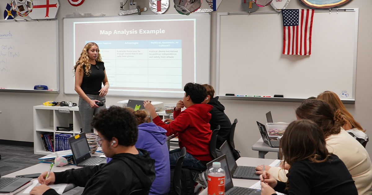A teacher speaking in front of a projector. Around her is a group of students, seated at their desks.