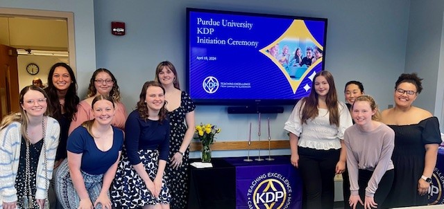 A group of KDP inductees standing in front of a welcome table. Behind them is a screen stating "Purdue University KDP Initiation Ceremony"