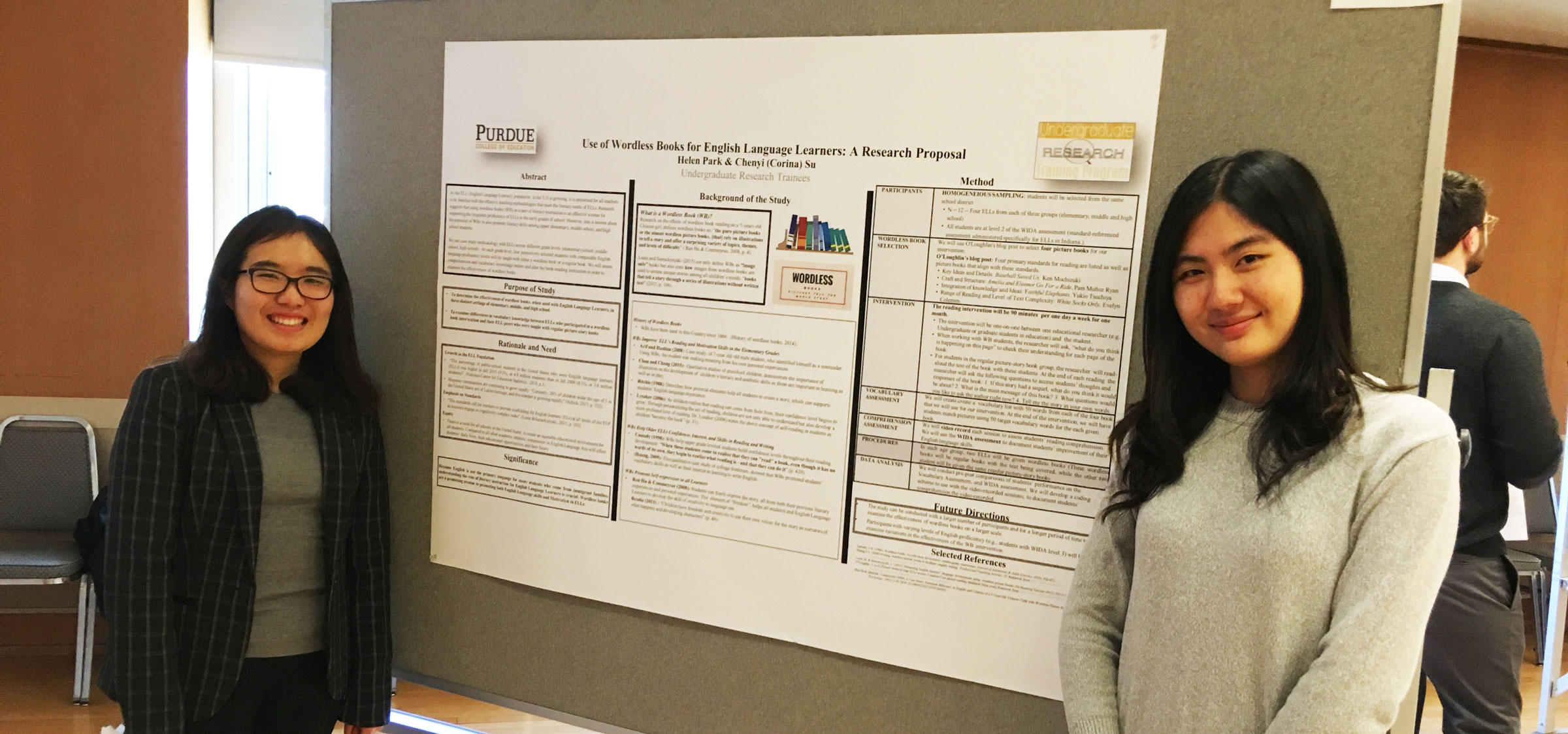 Two female Asian students stand in front of a poster presentation.