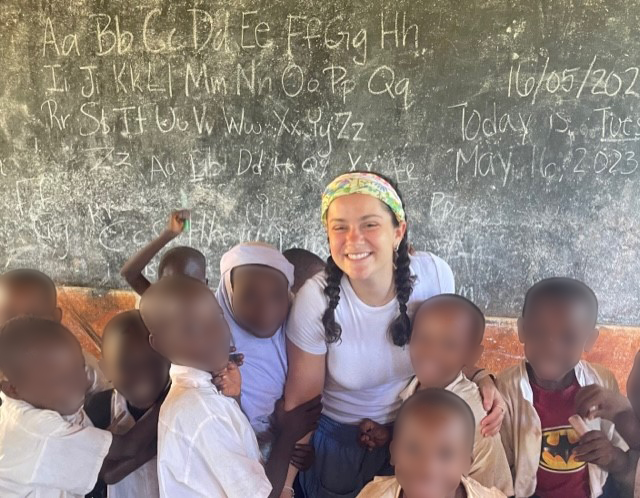 Lauren Zarras with a group of Tanzanian schoolchildren standing in front of a chalkboard.