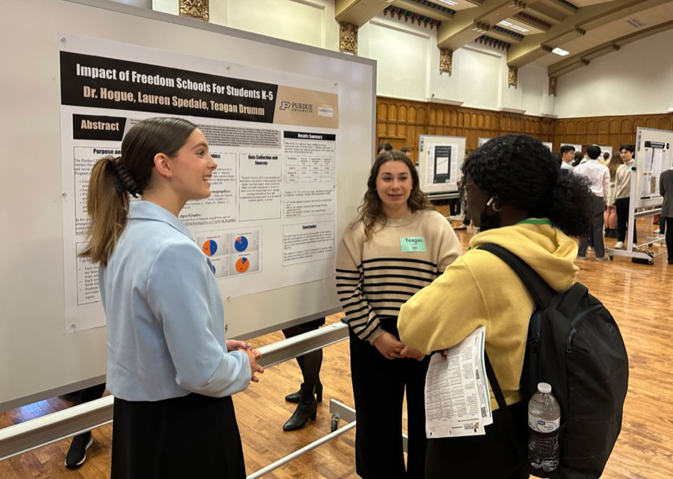 A group of female students stand discussing a poster presentation.