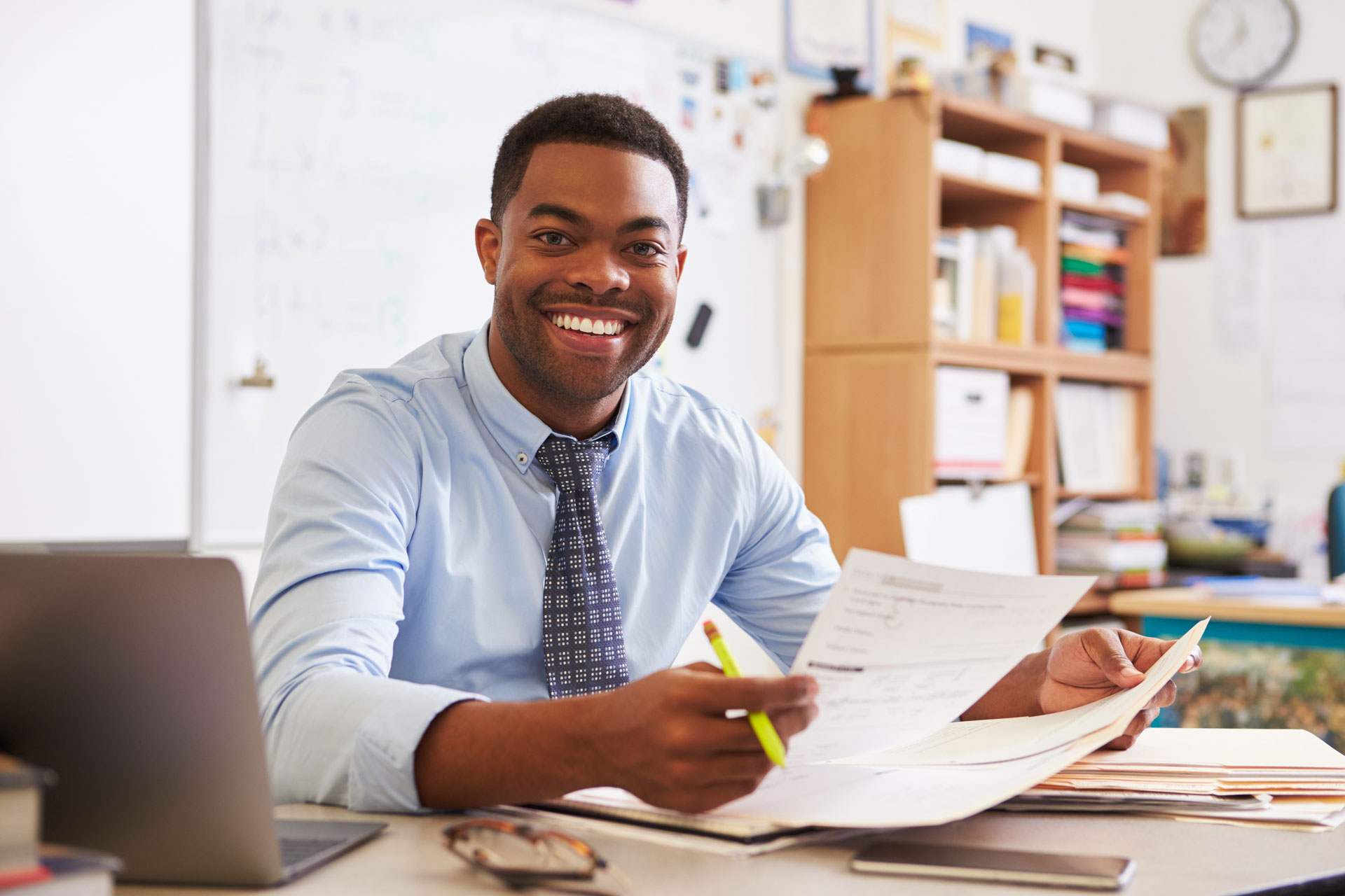 A black male teacher sits a desk in a classroom and grades papers
