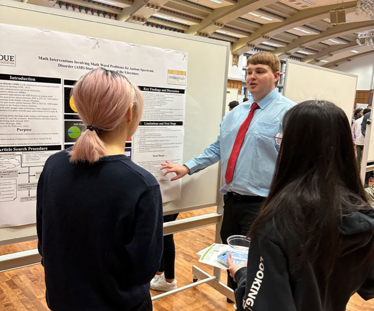 A white male student stands in front of a poster presentation with two onlookers.