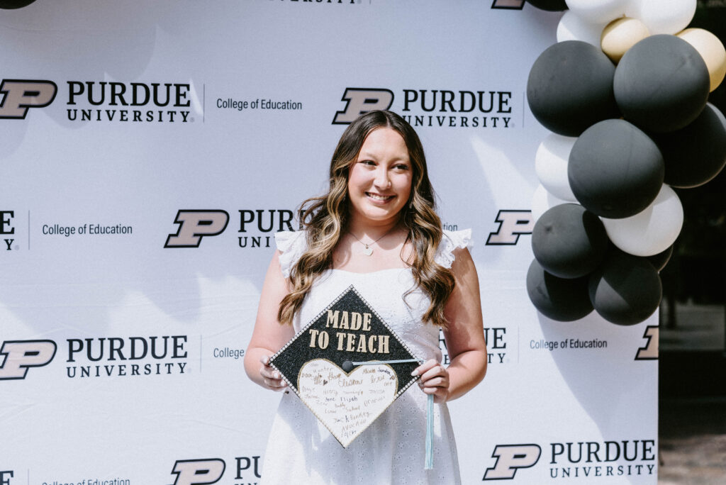 A College of Education graduate standing in front of a Purdue themed background. She is holding a graduation cap decorated with the words "Made to Teach".