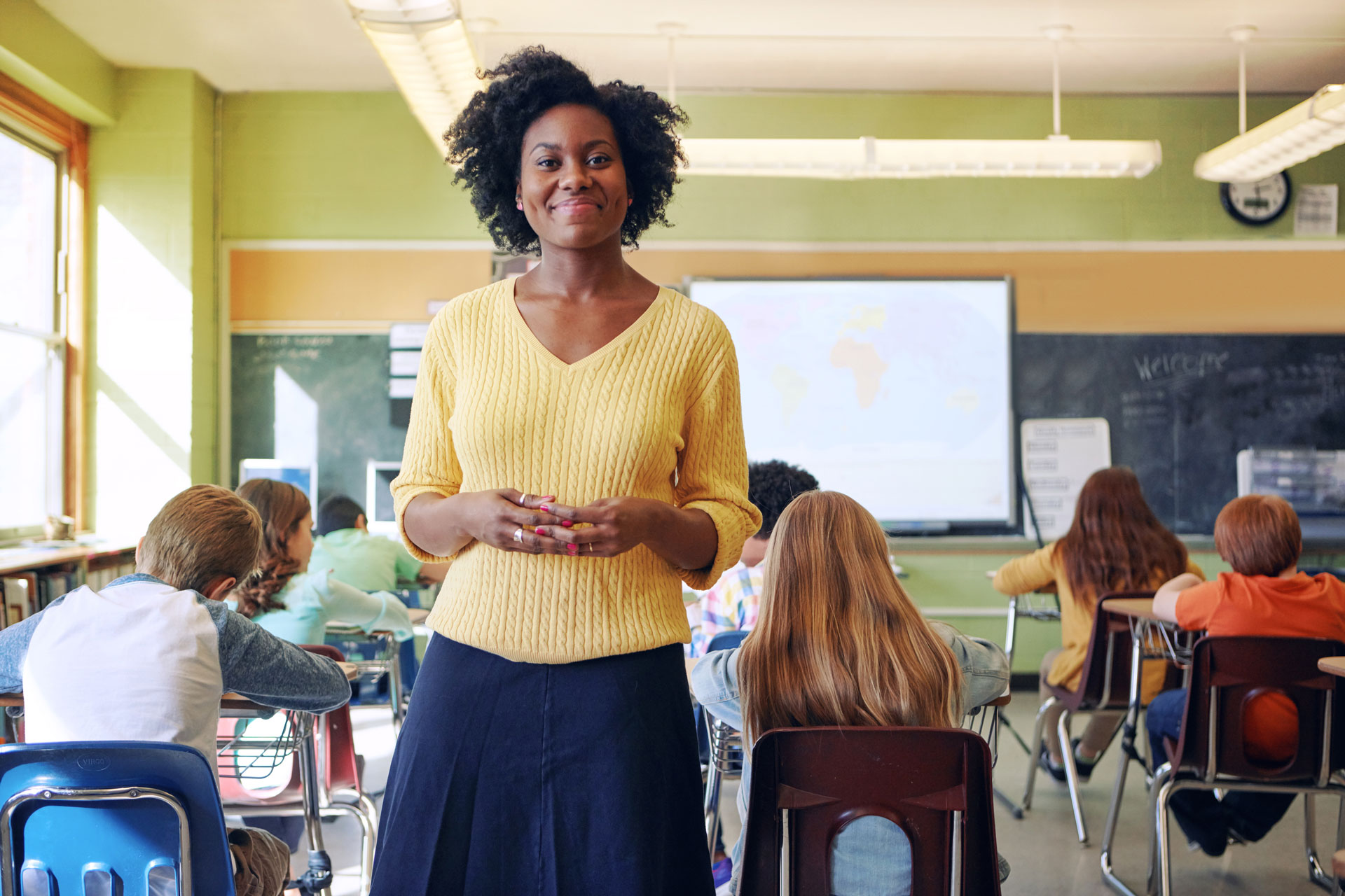 A black female teacher stands at the back of the classroom. A group of students are behind her.