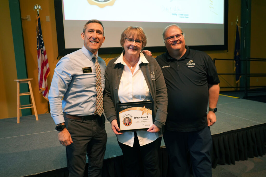 Rita Neidlinger holding a certificate for the 2023-2024 Bravo Award. Beside her are Interim Dean VanFossen and Wayne Wright