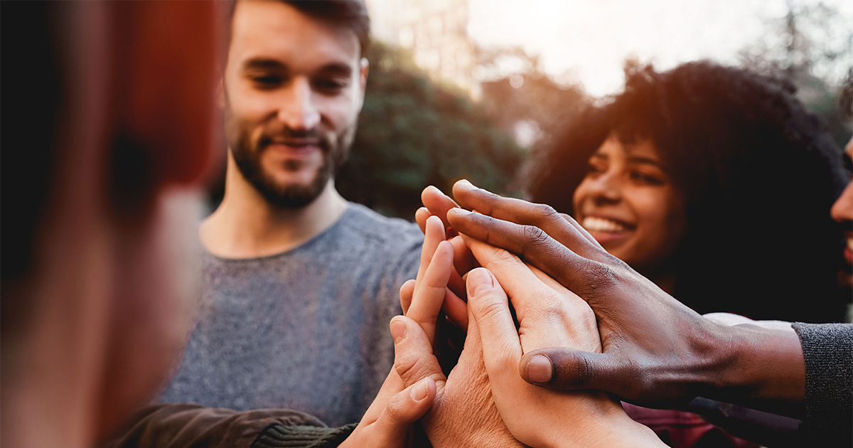 A group of people touch hands in the middle of a circle. The face of a white man and black woman are visible.