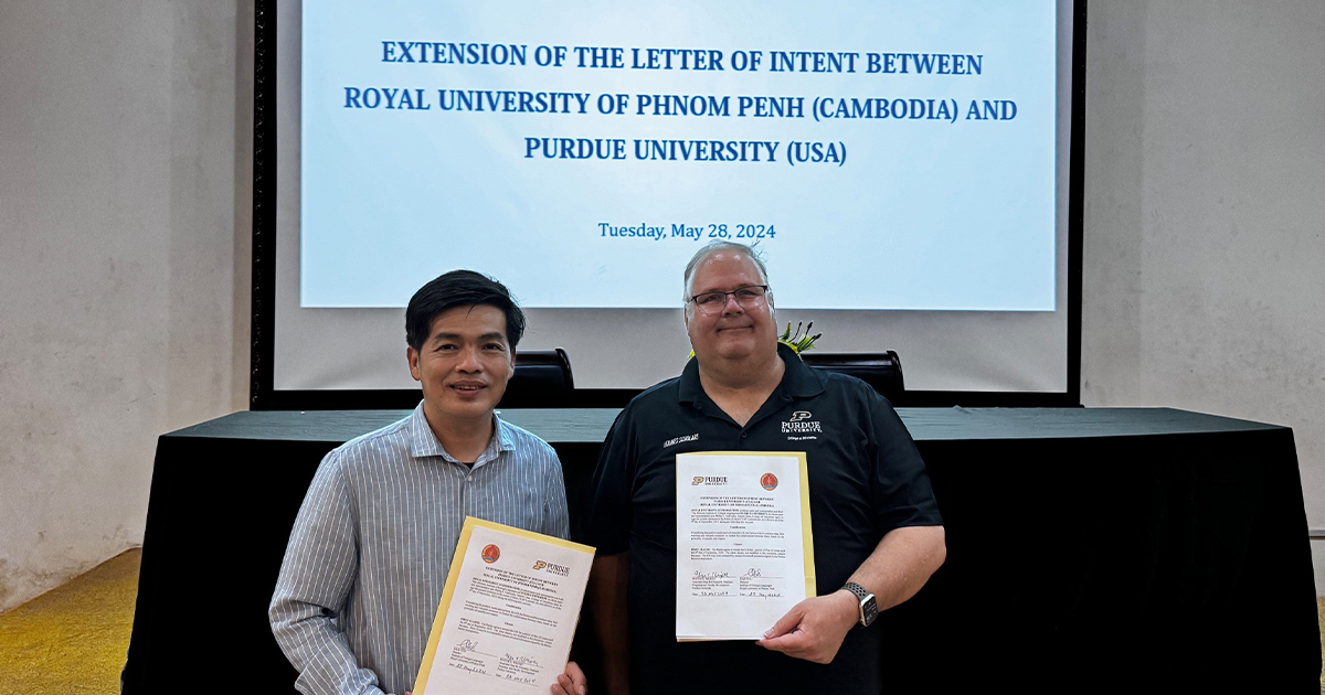 Wayne Wright and Tith Mab holding a signed document. Behind them is a PowerPoint presentation projected onto a white screen.