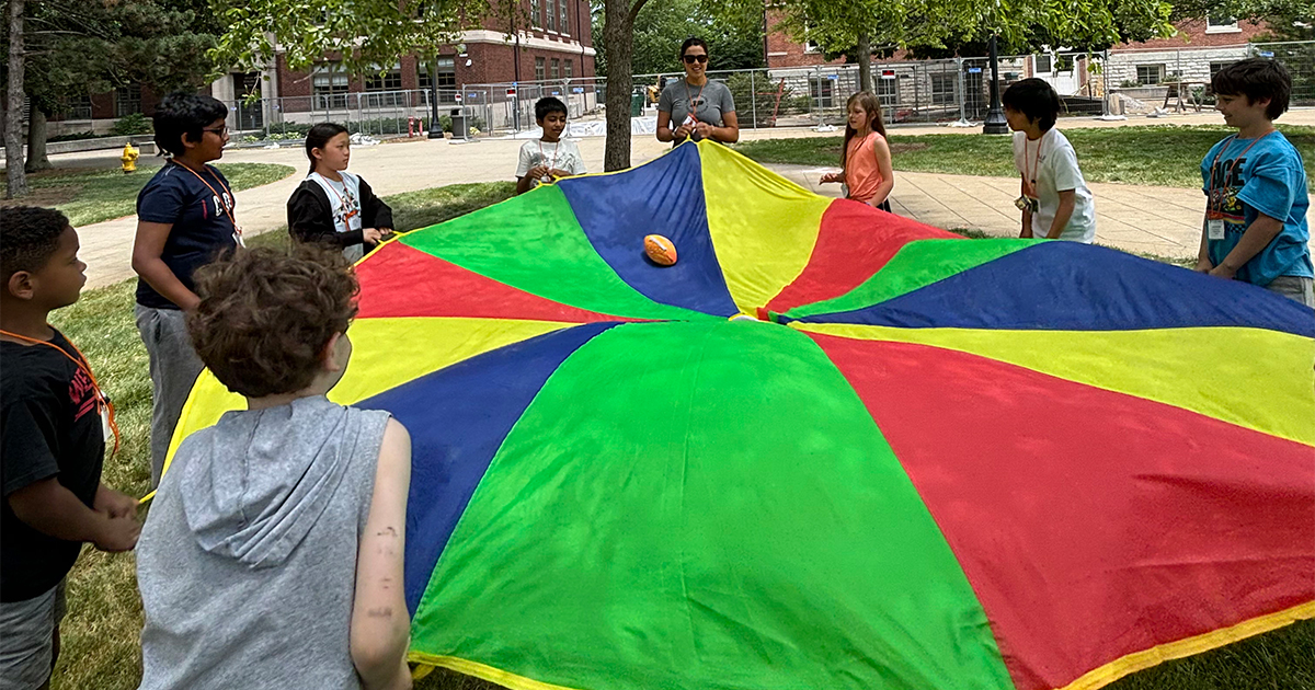 A group of GER2I students playing a teambuilding activity outside. Each student is holding a handle of a large circular colorful sheet.