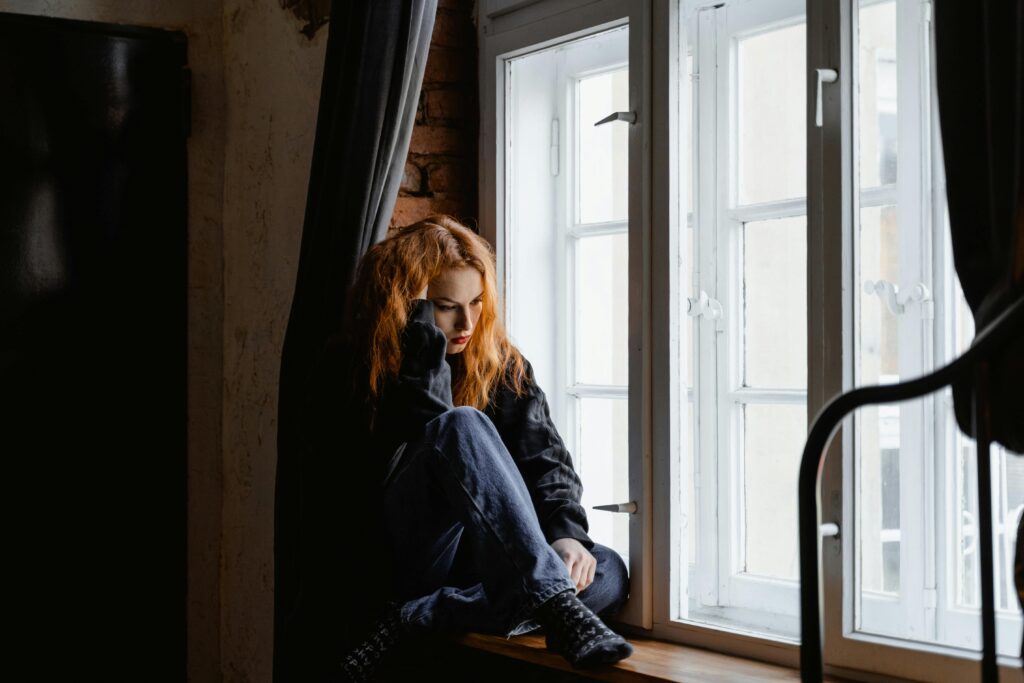 A young woman sitting on a window sill with a forlorn expression on her face.