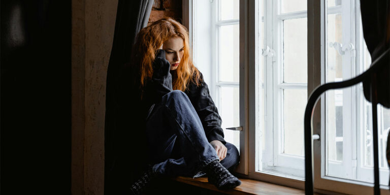 A young woman sitting on a window sill with a forlorn expression on her face.