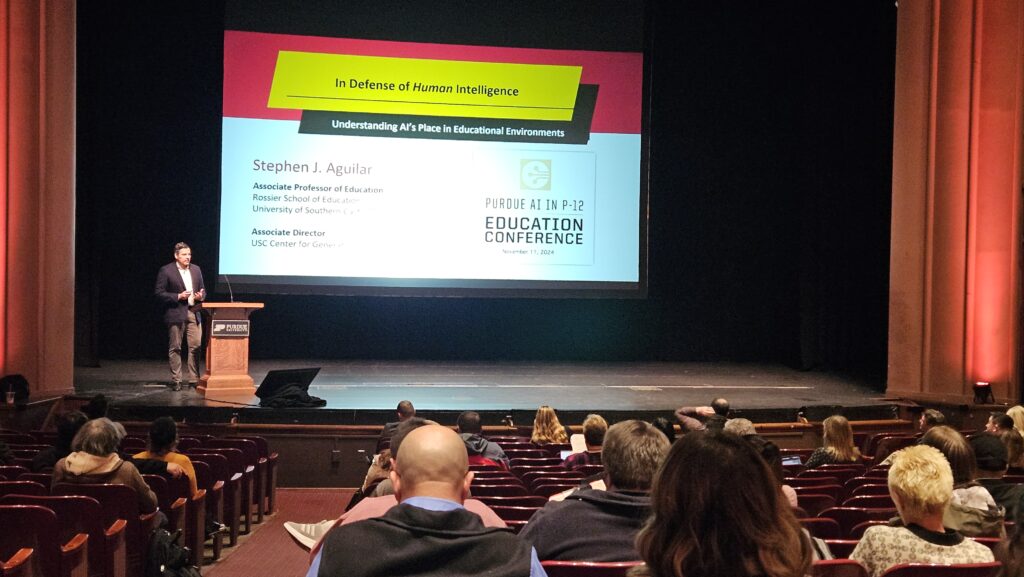 Stephen J. Aguilar standing at a podium in front of a projector screen featuring a presentation titled, "In Defense of Human Intelligence."