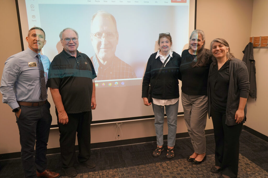 Phillip J. Van Fossen, James Greenan, JoAnn Phillion, Lynn Bryan, and Selcen Guzey all standing in front of a projection screen of Tim Newby, and smiling for a photo.