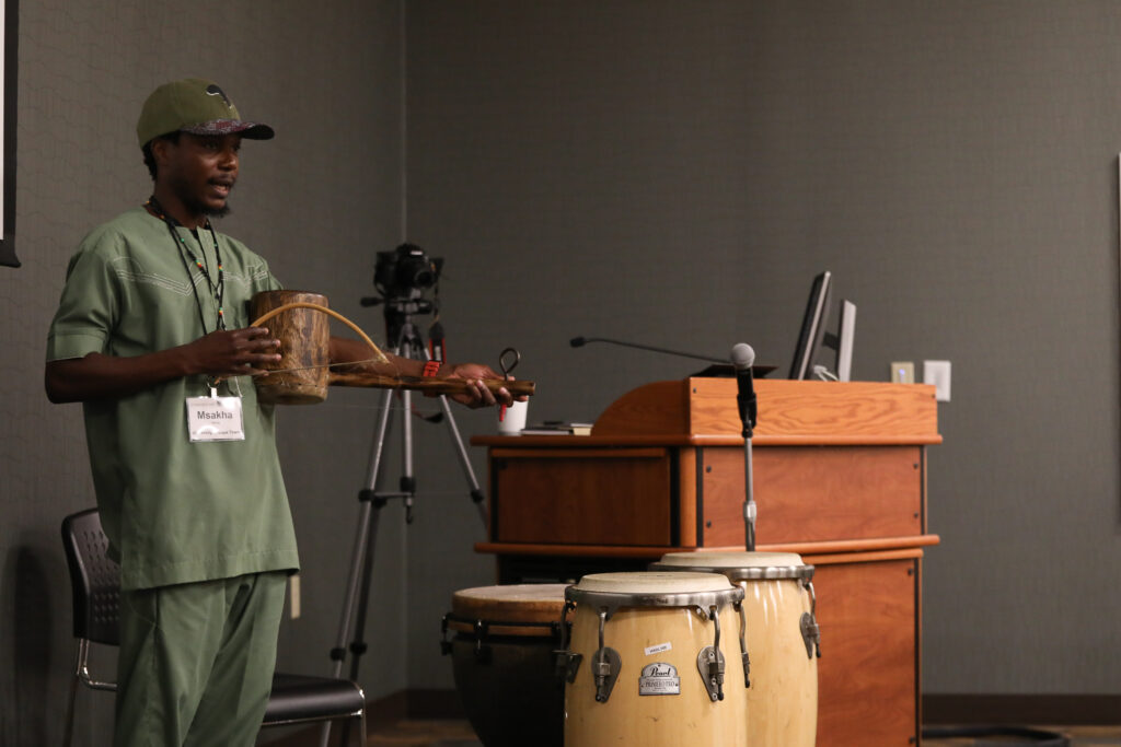 Msakha Mona at a podium, holding an instrument and speaking to ICLCLE attendees.