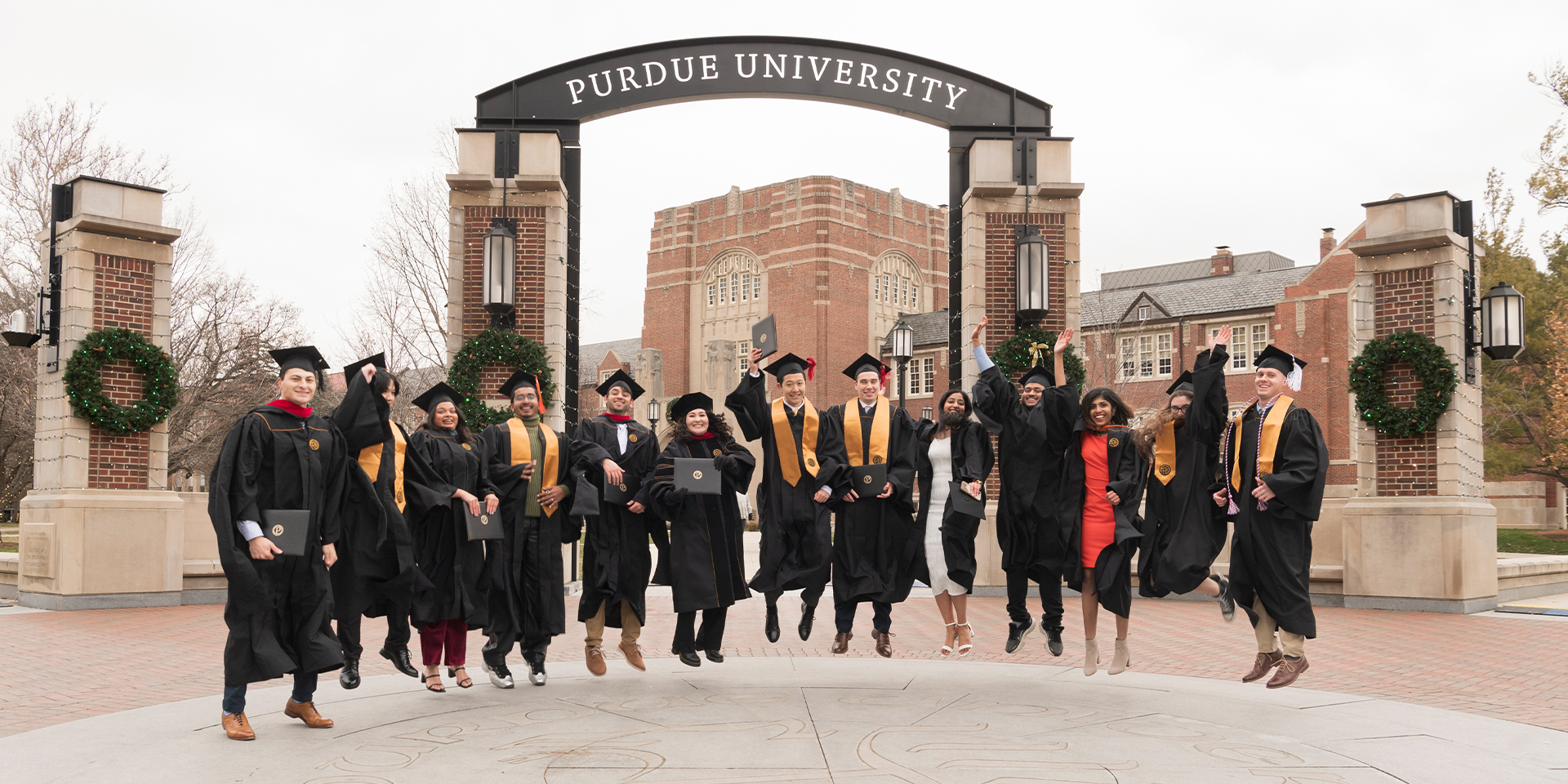 A group of Purdue graduates wearing their cap and gown and jumping in front of the Purdue arch.