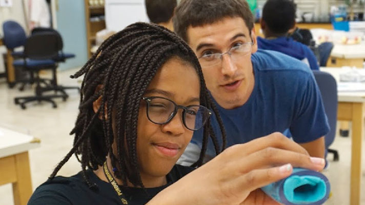 A young female student with glasses looks into a blue tube. Behind her, a camp counselor observes her.