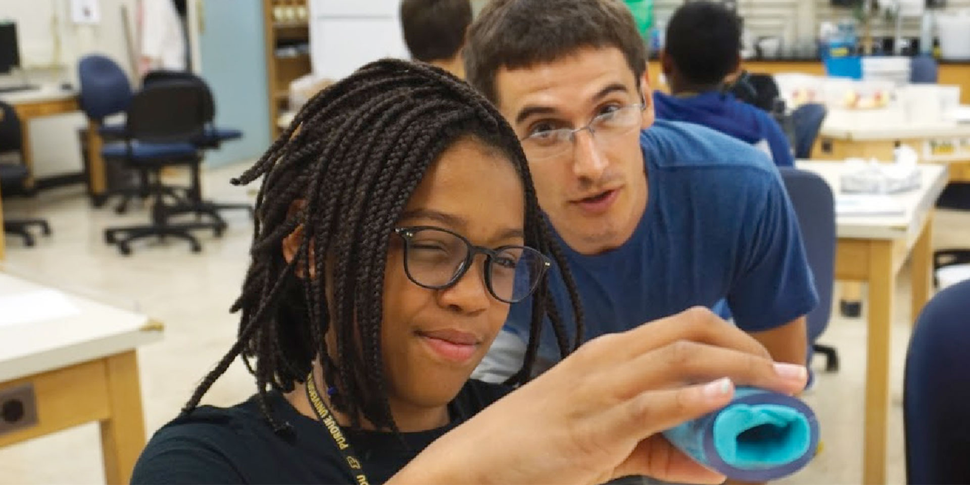 A young female student with glasses looks into a blue tube. Behind her, a camp counselor observes her.