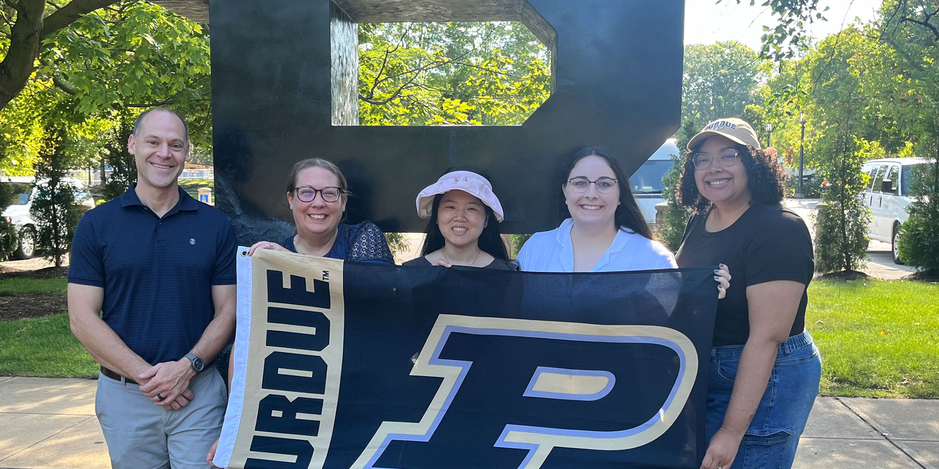 A group of 5 Purdue students holding a Purdue banner in front of a Block P statue outside.
