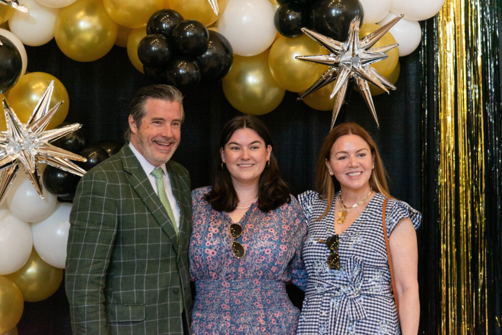 A newly graduated student and her family smiling for a photo in front of a black and gold backdrop.