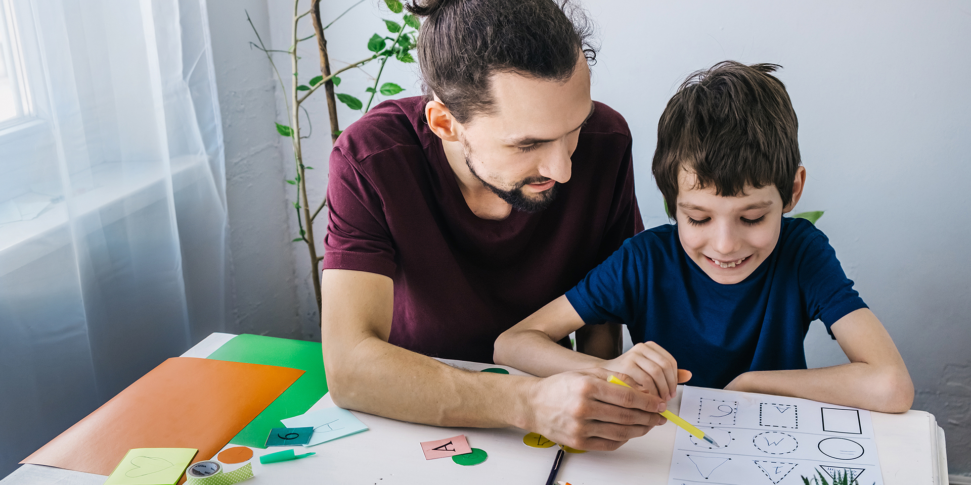 A male teacher in a burgundy shirt helping a young student wearing a blue shirt with his worksheet.