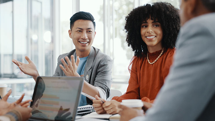 A team of professionals meeting in a conference room. An Asian man and African-American woman are featured as the subject. Also visible are notebooks, pens, and laptops.