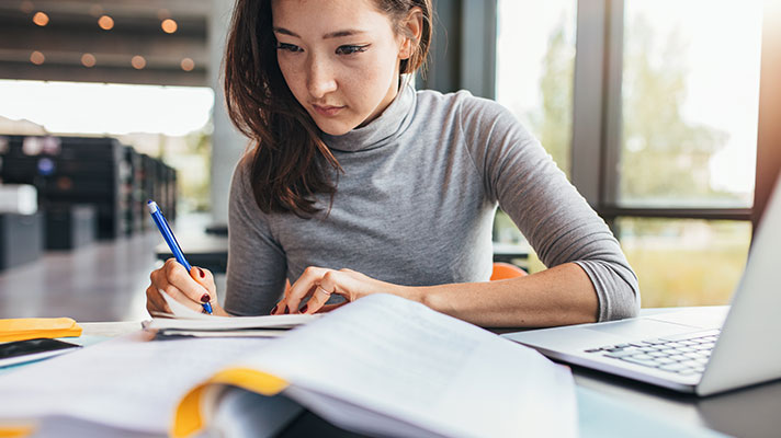 A student writes something in a notebook while looking at book and their laptop