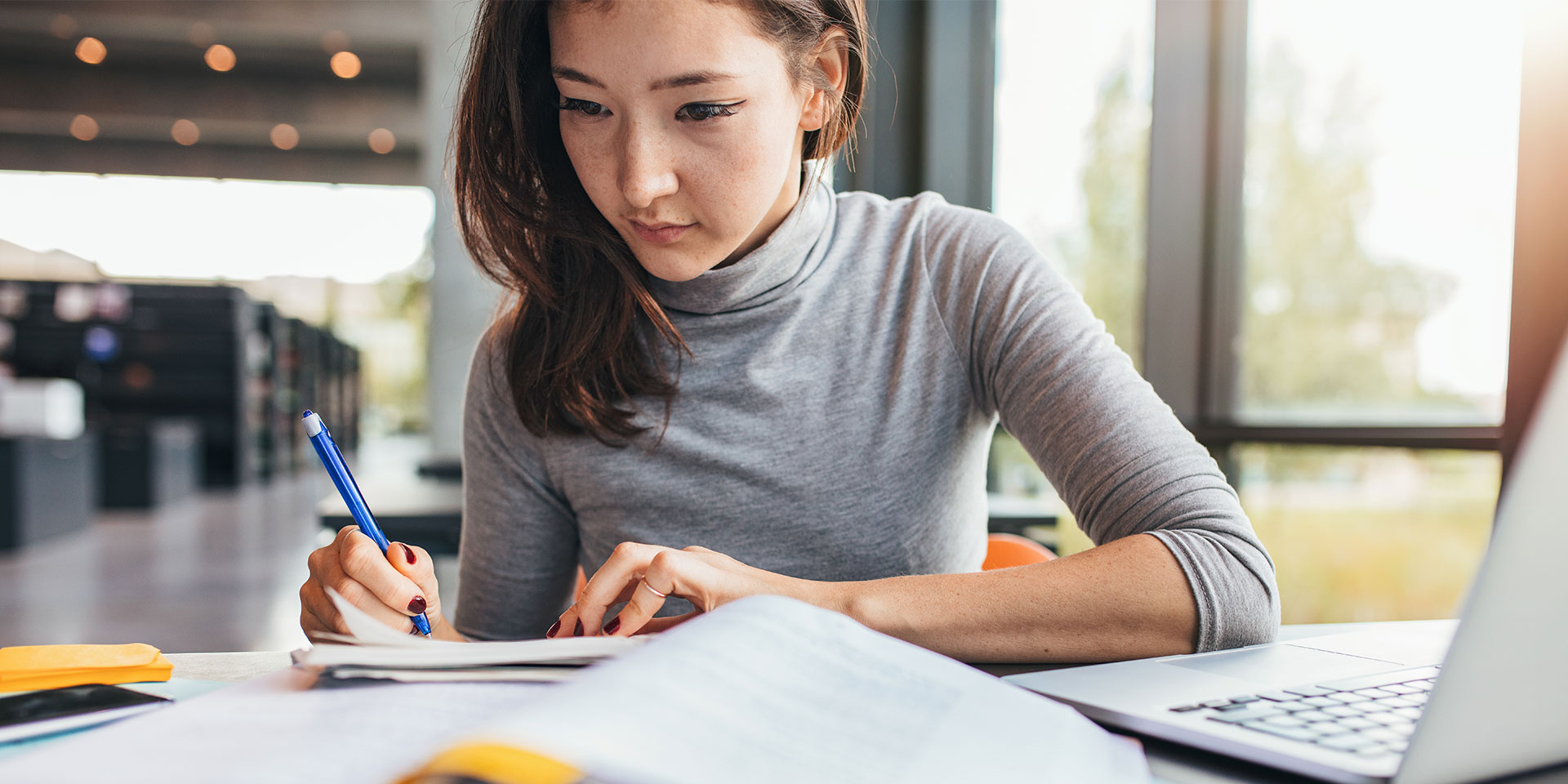 A student writes something in a notebook while looking at book and their laptop
