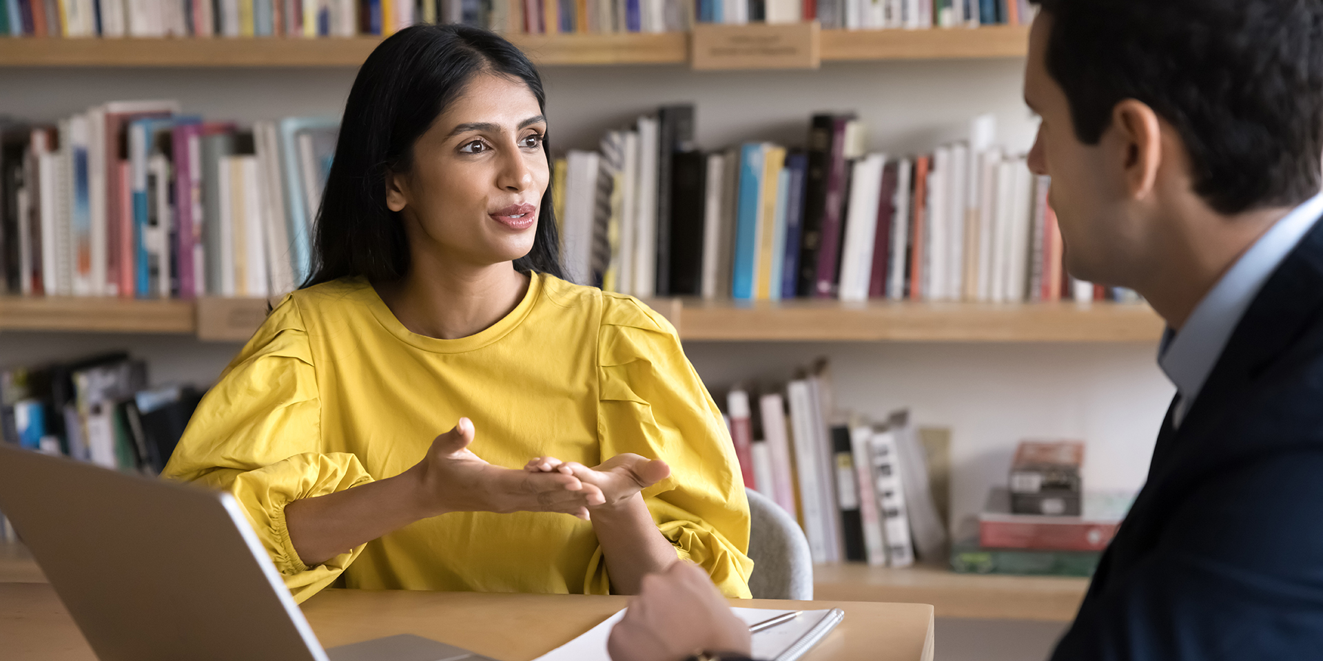 A young woman wearing a yellow shirt sits in a library. She is chatting with a man wearing a suit.