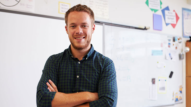 A Caucasian male teacher smiling in front of a white board with his arms crossed. He has short, reddish brown hair and is wearing a blue, checkered button-up shirt.