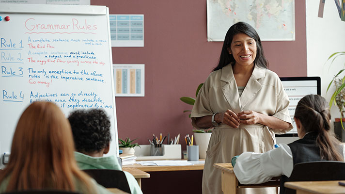 A female teacher stands in front of the classroom with a whiteboard featuring 