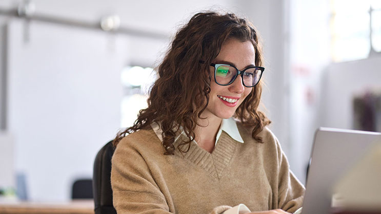 A young woman with glasses and wavy brown hair using a laptop.