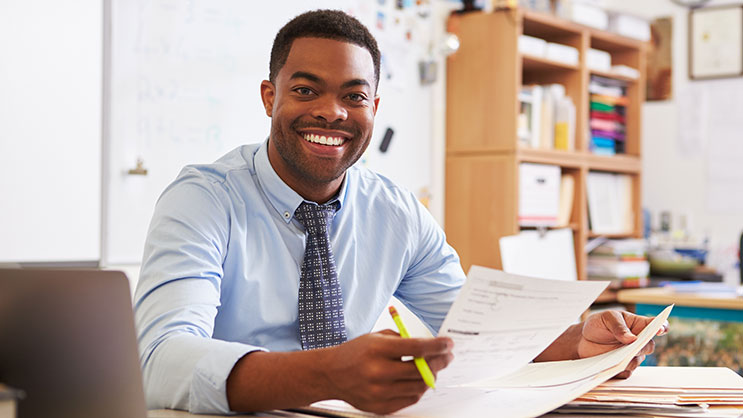 A young black teacher grades papers at a desk in a classroom. He is wearing a light blue dress shirt and a patterned navy tie. In his hands are a pencil and various papers.