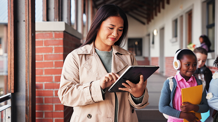 A young woman looking down at a tablet in her hands. She is wearing a light green knit sweater and a tan trench coat. Around her are grade school students walking to their next class.