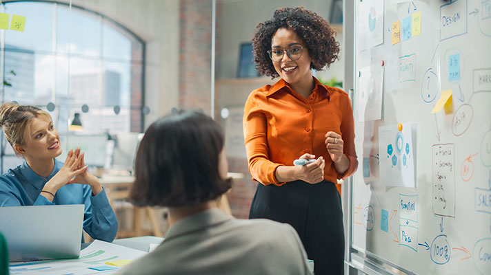 A woman stands in front of two of her colleagues in front of a whiteboard