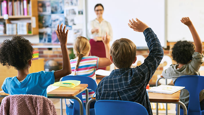 Students raise their hands while a teacher stands at the front of the classroom in the background